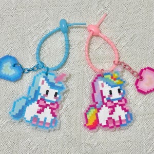 May include: Two pixelated unicorn keychains. One is blue with a blue heart charm, the other is pink with a pink heart charm. Both have a bow tie and colorful mane. The keychains are connected by a blue and pink loop.