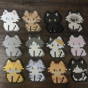 May include: A collection of twelve pixelated cat designs in various colors. The cats are in a sitting position, with details like blue eyes and patterned fur. The color palette includes white, black, gray, orange, and calico patterns.