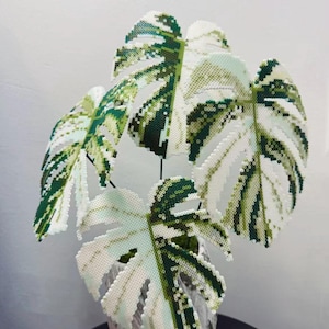 Op de afbeelding: Een decoratieve plantsculptuur gemaakt van kleine, gesmolten plastic kralen. De bladeren hebben een bont patroon van wit, lichtgroen en donkergroen, wat een Monstera-plant nabootst. De sculptuur staat in een pot met een textuur.