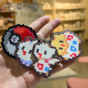 May include: Pixelated keychains featuring a red and white Pokeball, and three different Pokemon characters. The keychains are in a variety of colors, including red, white, yellow, and blue. The keychains are attached to a silver keyring.