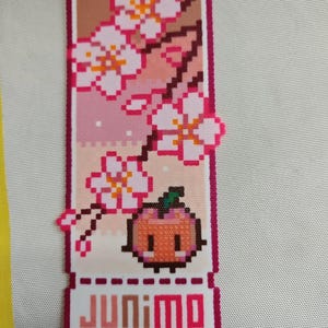 Op de afbeelding: Een pixelachtige boekenlegger met een kersenbloesemontwerp met roze en witte bloemen op een gradiënt achtergrond. Een kleine oranje vrucht met een groene steel staat onder de bloemen. Het woord "JUNIMO" staat onderaan.