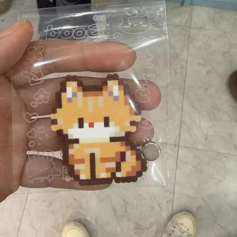 May include: A pixelated orange cat magnet in a clear plastic bag. The cat is in a sitting position with a white chest and black details. The bag has a cat-themed design.