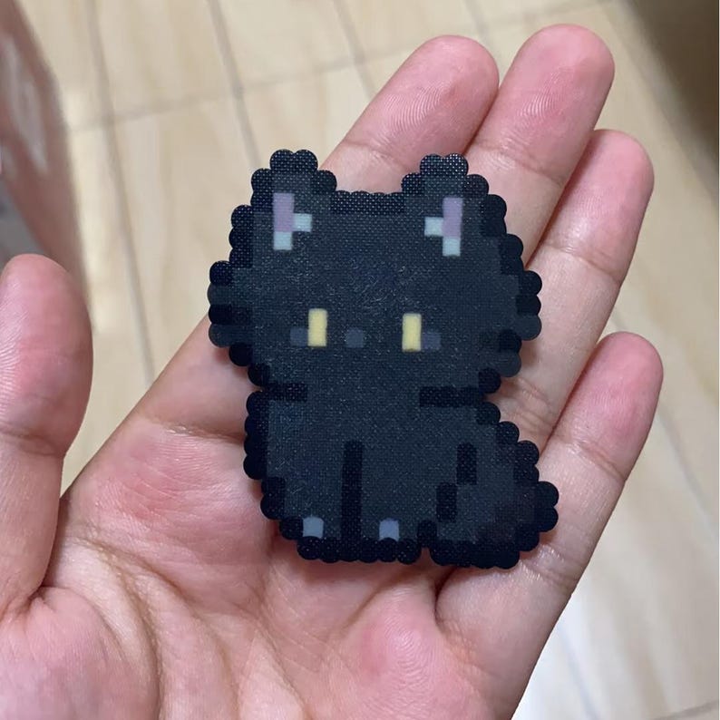 May include: A black pixel art cat sticker with yellow eyes and purple accents on the ears. The cat is sitting, and the sticker is held in a hand.