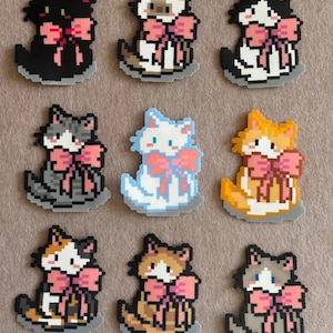May include: Nine pixelated cat-shaped charms in various colors, including black, white, gray, blue, and orange. Each cat has a pink bow and sits on a gray base. The charms are likely for crafting or jewelry making.