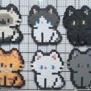 May include: Six pixelated cat-shaped patches in various colors: tan, black, orange, white, and gray. Each cat has a unique color scheme and facial features, with small eyes and a sitting pose. The patches are arranged on a grid.