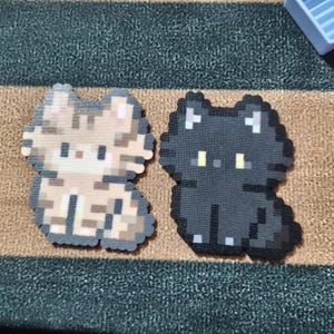 May include: Two pixel art cat figures. One is tan and gray, the other is black. Both are in a sitting position with small, square pixel details. The cats have small, yellow eyes and are on a striped surface.