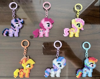 May include: Six pony-shaped keychains crafted from colorful plastic fuse beads. Each figure features a unique color palette including purple, pink, yellow, orange, white, and blue, all attached to matching plastic circular key rings.