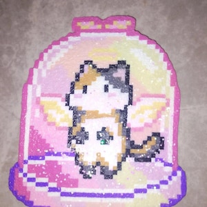 May include: A pixelated cat in a jar design. The cat is white, tan, and gray with small wings. The jar is pink, purple, and yellow with glitter accents. The design is made of small, colorful beads.