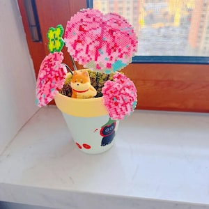 May include: A decorative flower pot with a light blue and yellow design, featuring a Shiba Inu figurine and pink and green beaded flowers. The pot has a cartoon cat illustration and sits on a white windowsill.