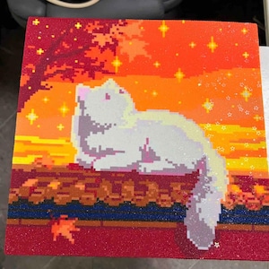 May include: A square wall art piece featuring a pixel art style white cat resting on a dark surface. The background displays an orange and red sunset sky with a tree silhouette and glowing yellow stars. The entire surface has a glittery texture.