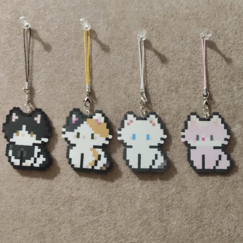 May include: Four pixelated cat charms in black, calico, white, and pink. Each charm has a different colored string and a silver clasp. The charms are hanging against a neutral background.