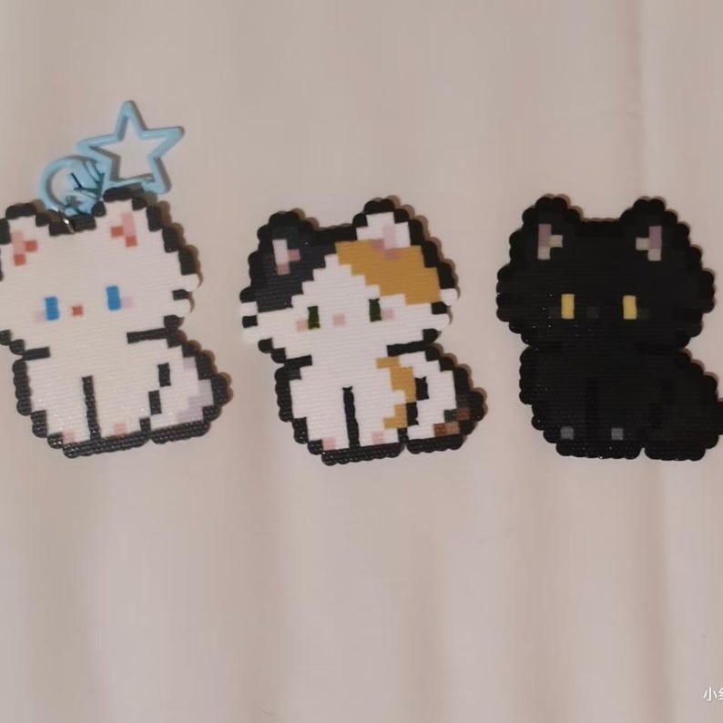 May include: Three pixel art cat charms. One white cat with blue eyes and a star charm, a calico cat with black, white, and orange, and a black cat with yellow eyes. The charms are made of small, colorful beads.