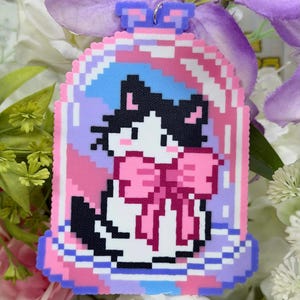 May include: A pixelated cat in a glass dome charm. The cat is black and white with a pink bow. The dome is surrounded by pink, blue, and purple pixelated details. The charm has a silver loop at the top for hanging.