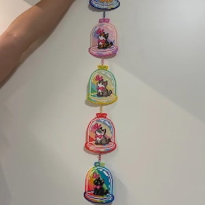 Op de afbeelding: Een reeks van vijf kleurrijke, gepixelde kattenornamenten, elk in een stolp. De katten dragen strikken en staan tegen een regenboogachtergrond. De stolpen zijn met gouden kettingen verbonden.