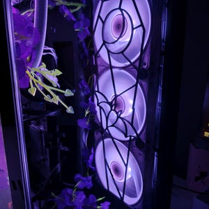 Puede incluir: Interior de una caja de ordenador con tres ventiladores iluminados, que emiten un suave brillo púrpura. Los ventiladores están enmarcados por un diseño geométrico. Flores púrpuras están dispuestas alrededor de los ventiladores y otros componentes.