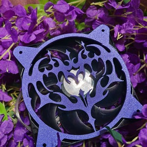 May include: A purple computer fan with a decorative, glittery, laser-cut design. The fan blades are black, and the fan is surrounded by purple flowers and green leaves. The fan has a silver centre.