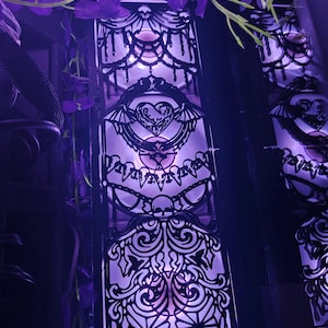May include: A decorative, illuminated panel with a gothic design. The panel features intricate black cutouts of hearts, wings, and ornate patterns. The panel is lit with a soft purple light, and is surrounded by purple flowers.
