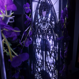 Puede incluir: Una caja de ordenador con un panel negro decorativo con un intrincado diseño gótico. Flores moradas caen en cascada alrededor de la caja, añadiendo un toque de color. El interior está iluminado con un suave resplandor púrpura.