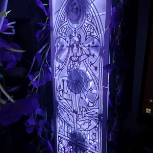 Peut inclure: Panneau de boîtier d'ordinateur avec un motif découpé au laser représentant un personnage de Sailor Moon dans un style vitrail. Le panneau est éclairé d'une douce lumière violette, et des fleurs violettes sont drapées autour.