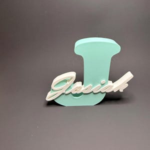 May include: A 3D-printed teal letter "J" with the name "Josiah" in white script. The name is layered over the letter. The background is a solid gray.