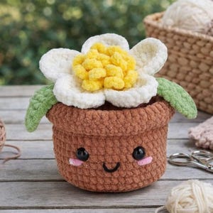 Op de afbeelding: Een gehaakte bloempot met een lachend gezicht, witte bloemblaadjes en een gele kern. De pot is bruin en de bladeren zijn groen. Garen en haakgereedschap staan op de achtergrond.