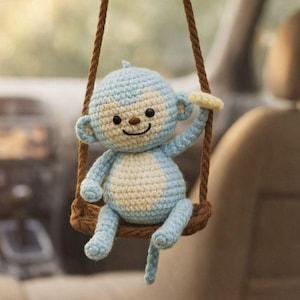 Może przedstawiać: Jasnoniebieska i kremowa szydełkowa małpka wisząca na brązowej huśtawce. Małpka ma uśmiechniętą twarz i trzyma żółtego banana. Huśtawka zawieszona jest na brązowej linie, urocza ozdoba do samochodu.