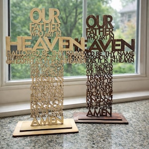 Könnte beinhalten: Zwei Holzkreuze mit dem Vaterunser eingraviert. Die Kreuze sind aus hellem und dunklem braunem Holz gefertigt und stehen auf rechteckigen Sockeln. Der Text enthält "Unser Vater" und "Amen".