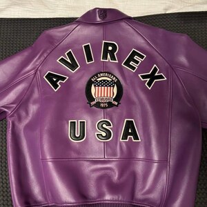 Op de afbeelding: Paarse leren jas met het woord "AVIREX" in witte blokletters op de rug. Daaronder een patch met de woorden "All Americans" en "1975". Het woord "USA" staat onder de patch.