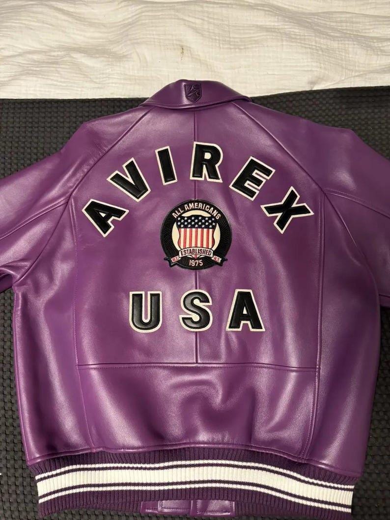 Op de afbeelding: Paarse leren jas met het woord "AVIREX" in witte blokletters op de rug. Daaronder een patch met de woorden "All Americans" en "1975". Het woord "USA" staat onder de patch.