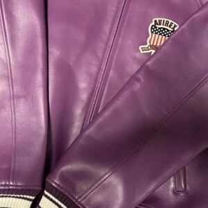 Op de afbeelding: Een close-up van een paarse leren jas met de "AVIREX" logopatch op de bovenborst. De jas heeft een geribbelde manchet met witte strepen. Het leer ziet er glad uit en heeft zichtbare stikdetails.