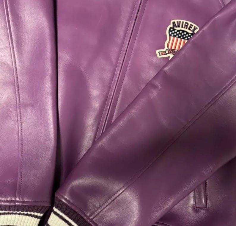 Op de afbeelding: Een close-up van een paarse leren jas met de "AVIREX" logopatch op de bovenborst. De jas heeft een geribbelde manchet met witte strepen. Het leer ziet er glad uit en heeft zichtbare stikdetails.