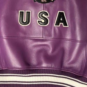 Op de afbeelding: Close-up van een paarse leren jas met het woord "USA" in witte letters met zwarte omlijning. De jas heeft een geribbelde zoom met witte en paarse strepen. Een patch is zichtbaar aan de bovenkant.