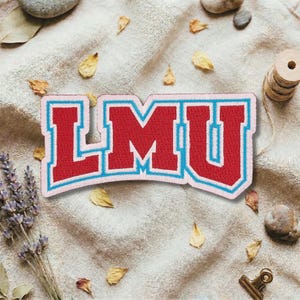 Op de afbeelding: Geborduurde patch met de letters "LMU" in een collegestijl. De rode letters hebben een lichtblauwe omtrek en een roze rand. De patch ligt op een beige stoffen achtergrond met gedroogde bladeren en lavendel takjes.