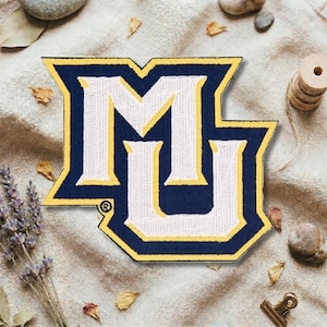 Puede incluir: Parche bordado con las letras "MU" en blanco, contorneadas en azul marino y amarillo. El parche, probablemente para una universidad o colegio, tiene un diseño colegial clásico. Está sobre un fondo de tela beige.