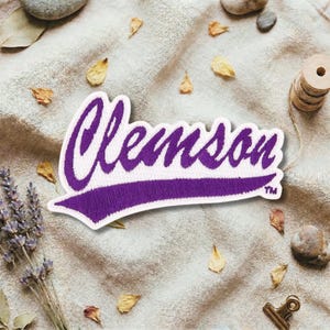 May include: A purple and white embroidered patch with the word "Clemson" in a script font. The patch has a white border and a purple tail. The patch is on a beige fabric background with dried leaves and lavender.