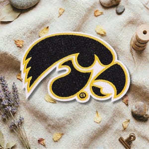Könnte beinhalten: Gestickter Aufnäher mit dem Logo der Iowa Hawkeyes der Universität. Der Aufnäher hat ein schwarz-goldenes Design mit weißem Rand. Das Logo ist ein stilisierter Falkenkopf mit dem Buchstaben "R" in einem Kreis.