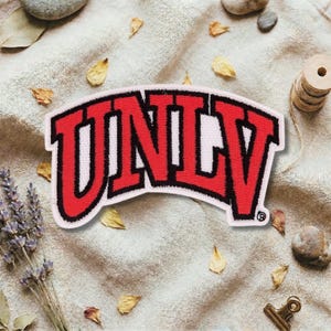 May include: A red and white UNLV patch with black trim. The patch is embroidered with the word "UNLV" in a bold, arched design. The patch is on a beige fabric background with dried leaves and lavender.