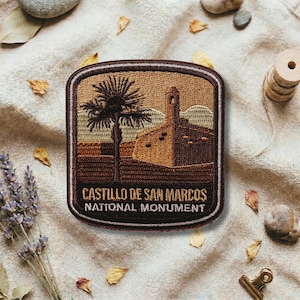 May include: Embroidered patch featuring the Castillo de San Marcos National Monument. The patch has a brown border and depicts a palm tree and a building with a tower in shades of brown and tan. Text reads "Castillo de San Marcos National Monument."