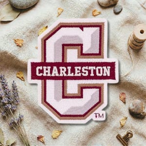 Könnte beinhalten: Gestickter Aufnäher mit einem großen Buchstaben "C" in Weiß, Hellgrau und Burgunderrot, mit dem Wort "CHARLESTON" in Burgunderrot. Der Aufnäher hat einen weißen Rand und ein "TM"-Symbol. Der Aufnäher befindet sich auf einer strukturierten, neutralfarbenen Oberfläche.