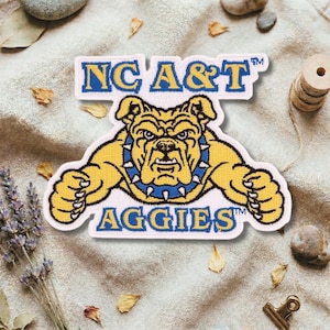 Może przedstawiać: Haftowana naszywka z logo NC A&T Aggies. Naszywka ma białe tło z napisem "NC A&T" w kolorze niebieskim i złotym. Żółty buldog z niebieską obrożą znajduje się na środku, a pod nim napis "AGGIES".