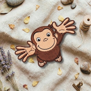 Op de afbeelding: Geborduurde patch met een lachende bruine aap met uitgestrekte armen. De patch heeft een bruin gezicht en handen, met zwarte contouren. De patch ligt op een beige stoffen achtergrond met gedroogde bladeren en lavendel takjes.