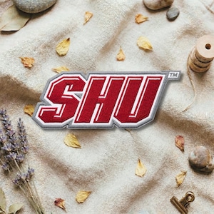 May include: A rectangular patch with the letters "SHU" in red with a white outline and a gray border. The patch is on a beige fabric surface, surrounded by dried leaves, lavender, and small stones.