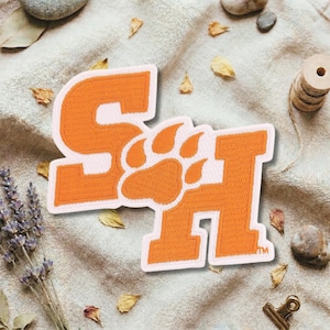 May include: An orange and white embroidered patch featuring the letters "S" and "H" with a paw print in the center. The patch is on a beige fabric background with dried leaves and lavender sprigs.