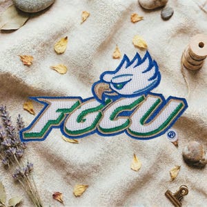 May include: Embroidered patch featuring the letters "FGCU" in white, green, and blue, with a stylized eagle head logo. The patch is on a beige fabric background, surrounded by dried leaves and small stones.