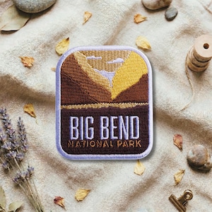 Puede incluir: Parche bordado con un diseño de paisaje y el texto "BIG BEND NATIONAL PARK". El parche es rectangular con esquinas redondeadas y un borde blanco. El diseño presenta tonos marrones, amarillos y blancos, que representan montañas y un cielo.