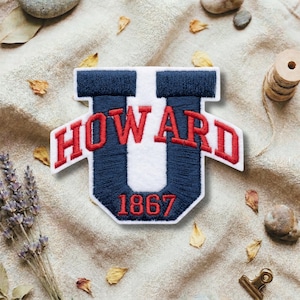 Puede incluir: Un parche bordado azul marino y blanco en forma de "U" con la palabra "HOWARD" en letras rojas. El número "1867" también es rojo. El parche está sobre un fondo de tela beige.