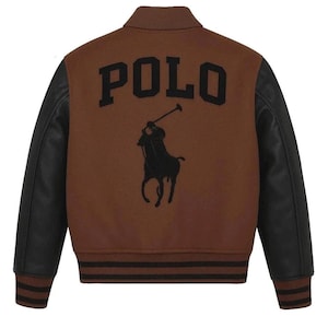 May include: Brown varsity jacket with black leather sleeves. The back features the word "POLO" in large black letters above a black silhouette of a polo player on a horse. The jacket has black and brown striped trim at the cuffs and waist.