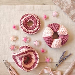 Puede incluir: Tres coleteros de ganchillo en tonos rosa y malva, con patrones de rayas y bloques de color. El conjunto incluye varios pequeños abalorios de plástico translúcido en forma de estrella y corazón, junto con dos ganchillos de metal.