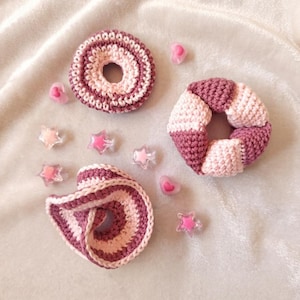 May include: Three crochet hair scrunchies in shades of pink and mauve, featuring varied striped and color-blocked patterns. The set is accompanied by several small, translucent plastic embellishments shaped like hearts and stars in pink tones.