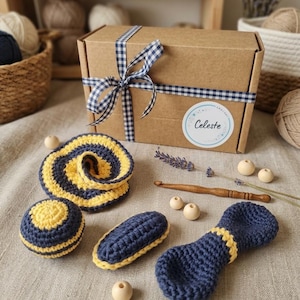 May include: A brown cardboard gift box tied with a blue and white gingham ribbon, featuring a circular label with the text Celeste. Included are several navy blue and yellow crocheted items, a wooden crochet hook, and small wooden beads.